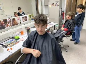 Ein Junge sitzt im Schminkraum der Maskenabteilung vor einem großen Spiegel. Er trägt eine aufgeschminkte Wunde im Gesicht. 