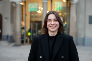 Portrait von Christiane Paul vor dem Shop des Berliner Ensembles. Sie trägt einen dunklen Mantel und lacht herzlich in die Kamera. 