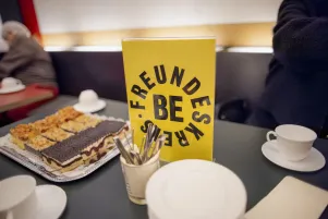 Schnappschuss: Auf einem Kantinentisch stehen einige Teller und Kuchen, daneben ist ein gelbes Schild zu erkennen, das auf eine exklusive Veranstaltung des Freundeskreises verweist. 