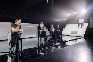 Schauspieler:innen auf einer hell ausgeleuchteten Bühne mit weißen Stufen und reflektierendem Boden.