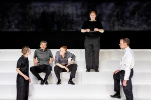 Mehrere Schauspieler:innen auf gestufter Bühne; eine Spielerin steht erhöht, während andere im Vordergrund diskutieren.