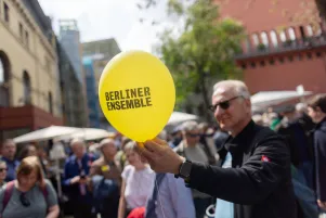 Ein Mann hält einen gelben Luftballon in der Hand, auf dem das Logo des Berliner Ensembles zu erkennen ist. 