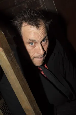 Nahaufnahme von Marc Hosemann aus erhöhter Position. Er trägt schwarzen Anzug und Krawatte, darunter ein rotes Hemd und schaut nach oben in die Kamera. Er scheint an einem Holzbalken zu lehnen. Der HIntergrund ist schwarz.