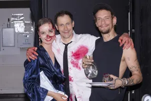 Clara De Pin und Marc Hosemann posieren mit einem Mitarbeiter aus der Requisite in ihren mit Kunstblut befleckten Bühnenkostümen. Alle schauen in die Kamera. Clara De Pin hat auch im Gesicht Kunstblut, Marc Hosemann an den Händen. Sie trägt einen blau-glänzenden Morgenmantel, Hosemann ein weißes Hemd und schwarze Krawatte.