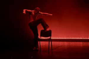 Darsteller in weiter Jeans auf einer, bis auf einen Stuhl, leeren Bühne. Die Bühne ist in rotes Licht getaucht, im Hintergrund sind LED-Leuchten zu erkennen. Mit großer Bewegung und den Händen zu Fäusten geballt, scheint er gerade auf den Stuhl zu steigen.