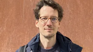Portraitaufnahme von Christian Staas in Mantel vor einer braunen Wand. Er trägt Brille und schaut in die Kamera.
