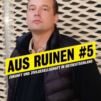 Autor und Host Hendrik Bolz lehnt an der Aussenwand des Neuen Hauses, er legt den Kopf etwas in den Nacken und schaut herausfordernd in die Kamera. 