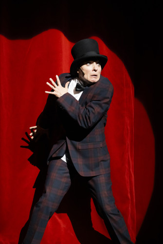 14)	Person in kariertem Anzug und Zylinder steht vor rotem Vorhang, hebt eine Hand mit gespreizten Fingern und hat eine Fliege an.