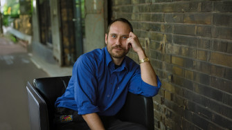 Portrait von Ensemblemitglied Sebastian Zimmler. Er sitzt in einem schwarzen Ledersessel vor einer Backsteinwand, trägt ein blaues Hemd, stützt sich auf seinen linken Arm auf und schaut direkt in die Kamera. 