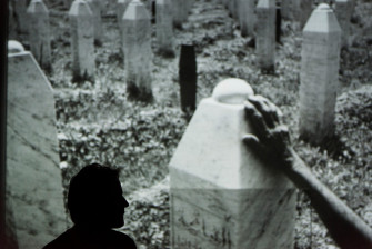Inszenierungsfoto von Srebrenica, links im Bild die Umrisse von Jens Harzer vor einer Projektion eine Fotos im Hintergrund. Auf dem Foto sind Grabsteine in Form von Stelen zu erkennen sowie eine Hand, die einen dieser berührt. 