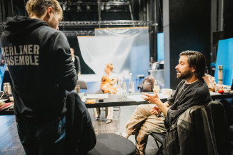 Regisseur Antú Romero Nunes sitzt am Regietisch und unterhält sich mit einem Kollegen, dessen schwarzer Hoodie mit der Aufschrift "Berliner Ensemble" der Kamera zugewandt ist. 