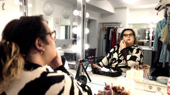 Der Creator Timur sitzt im Schminkraum vor einem großen Spiegel mit Glühbirnen. Links im Bild ist seine Schulter zu erkennen, das Foto zeigt aber sein Spiegelbild. Er ist bereits extravagant geschminkt und hält einen Pinsel in der Hand, mit dem er sich bepudert. 