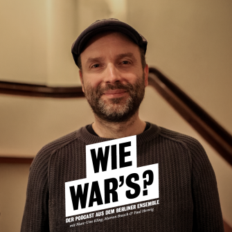 Portrait von Marc-Uwe Kling im Treppenaufgang zum ersten Rang im Großen Haus. Er trägt einen grau-braunen Pullover und eine Schiebermütze. Er lächelt direkt in die Kamera. 