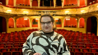 Timur steht auf der Bühne des Großen Hauses, im Hintergrund ist der  hell erleuchtete Zuschauerraum zu erkennen. Er schaut direkt in die Kamera. 