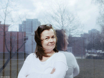 Portrait der Autorin Katja Oskamp vor einer spiegelnden Glasfläche. Es sind Hochhäuser zu erkennen. Sie trägt ein weißes Oberteil, verschränkt die Arme und schaut seitlich in die Kamera. 