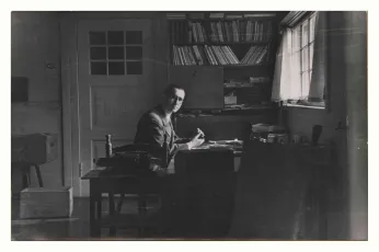 Bertolt Brecht an seinem Schreibtisch in seiner Wohnung, im Hintergrund ein Bücherregal.