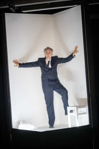 Jens Harzer im Bühnenbild. Er befindet sich in einem schmalen hohen und weißen Kasten, er trägt einen dunklen Anzug. 