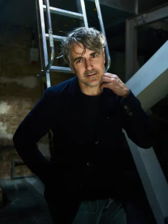 Schauspieler Jens Harzer steht in schwarzem Outfit vor einer Leiter unter der Bühne. Er schaut in die Kamera, hat eine Hand am Hals und wird von oben beleuchtet. 