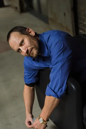 Portrait von Ensemblemitglied Sebastian Zimmler. Er hängt sich über die Lehne eines schwarzen Ledersessels vor einer Backsteinwand, trägt ein blaues Hemd, stützt sich auf seinen linken Arm auf und schaut direkt in die Kamera. 