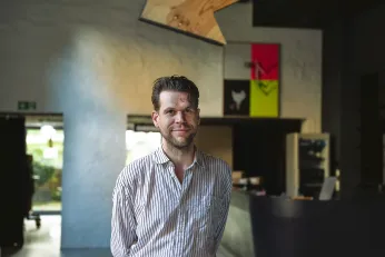 Ein Portrait des Künstlers Alex Wissel, er trägt ein weißes Hemd, schaut direkt in die Kamera und steht im Foyer des Neuen Hauses. Im Hintergrund sind schemenhaft einige seiner Arbeiten zu erkennen. 