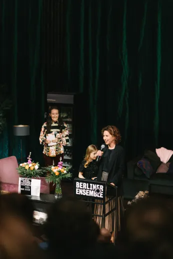 Tena Štivičić steht vor einem hohen Tisch mit dem Logo des Freundeskreises im Bühnenbild von "Die Verstreuten". Sie spricht in ein Mikrofon, neben ihr sind ihre Tochter und ein Vorstandsmitglied des Freundeskreises zu erkennen. 