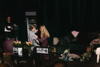 Amelie Willberg und Kathleen Morgeneyer stehen nah beieinander und lachen einander an. Um sie herum ist das Bühnenbild von "Die Verstreuten" zu erkennen. 