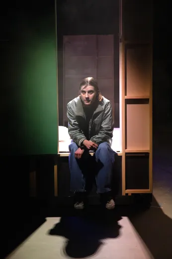 Amelie Willberg sitzt an der vorderen Kante des höher gestellten Bühnenbildes (Wohnzimmer). Sie trägt blaue Jeans und grau-grüne Bomberjacke, stützt sich mit ihren Unterarmen auf den Knien ab, Blick nach vorne gerichtet.