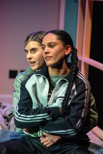 Nahaufnahme von Amelie Willberg und Amal Keller, die hintereinander aneinander umarmt sitzen. Im Hintergrund ist unscharf das Bühnenbild des Wohnzimmers zu erkennen.