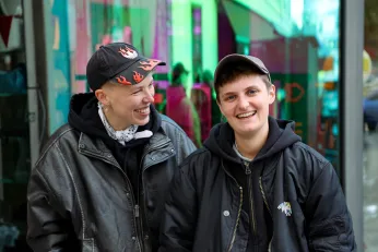 Leo Loreny Wyss und Jules Head stehen jeweils in schwarzen Bomberjacken und mit dunklen Caps lachend vor der bunt reflektierenden Glasfront des BE-Shops.