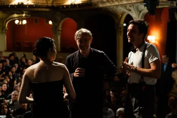 Behind the Scenes-Schnappschuss. Der Regisseur bespricht sich mit seinem Team, dabei stehen sie auf der Bühne des Berliner Ensembles, im Hintergrund ist ein gefüllter Saal zu erkennen.