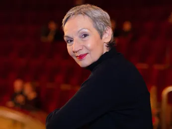 Ein Portrait von Moderatorin Marion Brasch im ersten Rang des Großen Hauses. Sie trägt einen schwarzen Rollkragenpullover und lehnt sich auf die Rangbrüstung. Dabei schaut sie über die Schulter direkt in die Kamera. 