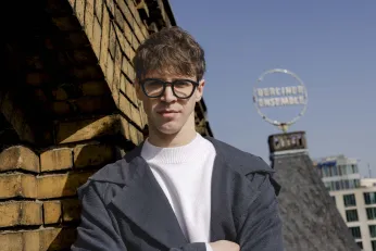 Portrait von Piotr Pacześniak auf dem Dach des Berliner Ensembles. Er lehnt an einer gelben Backsteinwand und schaut direkt in die Kamera. Im Hintergrund ist das Logo des BE zu erkennen. 
