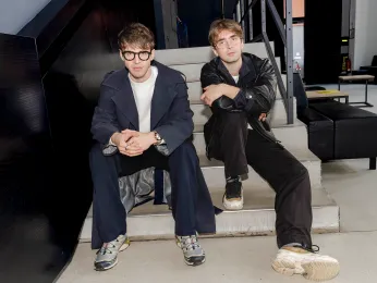 Luis August Krawen und Piotr Pacześniak sitzen in lockerer Pose auf der Treppe im Foyer des Neuen Hauses. Die Kamera befindet sich auf deren Augenhöhe, sie schauen direkt in die Kamera. 