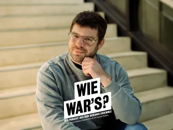 Portrait von Simon Dömer im Treppenhaus des Neuen Hauses. Er sitzt auf den Betonstufen, trägt Brille und ein blaugraues Oberteil. Das Kinn hat er linksseitig auf den gewinkelten linken Arm gestützt.