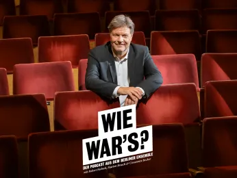 Robert Habeck sitzt auf den roten Samtsitzen im Zuschauerraum, die Unterarme hat er auf den Rückenlehnen der Vordersitze abgelegt. Er lächelt direkt in die Kamera. 