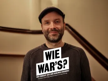 Portrait von Marc-Uwe Kling im  Treppenaufgang zum ersten Rang des Großen Hauses. Er trägt einen braunen Pullover, eine Cap und lächelt in die Kamera. 