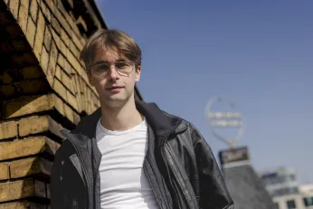 Portrait Luis August Krawen auf dem Dach des Berliner Ensembles, links ist eine gelbe Backsteinwand zu erkennen, im Hintergrund das Logo des BE.  Das Foto ist leicht von unten aufgenommen, er schaut direkt in die Kamera. 