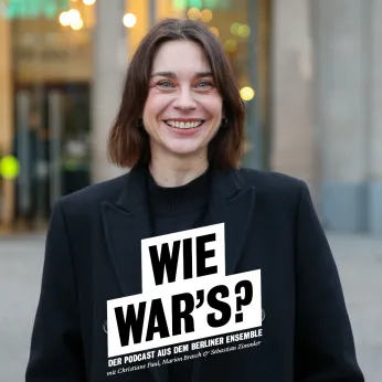 Portrait von Christiane Paul vor dem Berliner Ensemble, im Hintergrund ist der Shop zu erkennen. Sie trägt einen dunklen Mantel und lacht offen in die Kamera. 