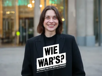 Portrait von Christiane Paul vor dem Berliner Ensemble. Im Hintergrund ist der Shop zu erkennen. Sie trägt einen dunklen Mantel und lacht offen in die Kamera. 