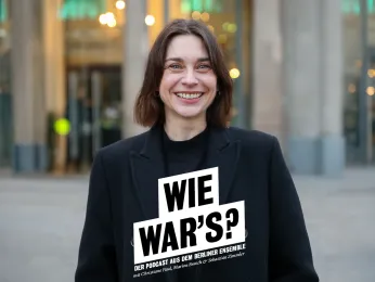 Portrait von Christiane Paul vor dem Berliner Ensemble, im Hintergrund ist der Shop zu erkennen. Sie trägt einen dunklen Mantel und lacht offen in die Kamera. 