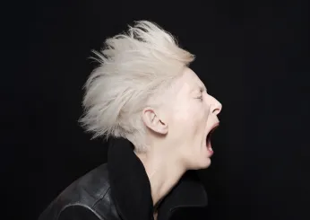 Aufnahme von Tilda Swinton im Seitenprofil. Der Hintergrund ist schwarz. Sie trägt eine schwarze Lederjacke mit Kragen, hat kurze platinblonde Haare und schreit.