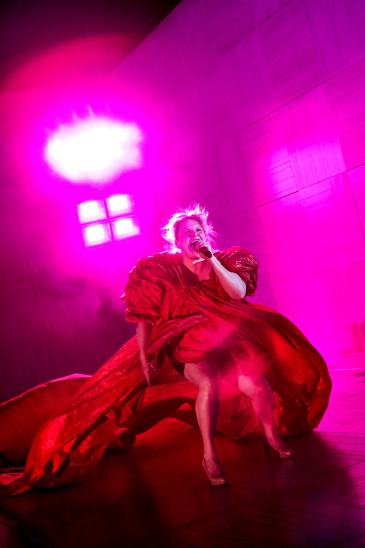 Frau in einem voluminösen roten Kleid singt mit Mikrofon auf einer Bühne mit pinker Beleuchtung.