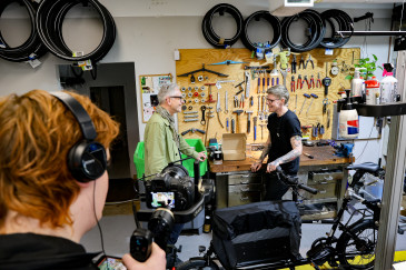 Behind the scenes: Oliver Reese und Jenny Andersen unterhalten sich vor der Werkbank, im linken BIldbereich ist der Kameramann samt Ausrüstung zu erkennen. 