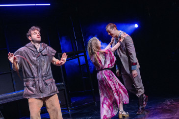 Szenenfoto "Mutti, was machst du da?" mit zwei Personen, die sich glücklich anschauen, dritte Person in Lederoberteil steht schulterzuckend daneben