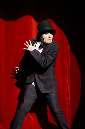 14)	Person in kariertem Anzug und Zylinder steht vor rotem Vorhang, hebt eine Hand mit gespreizten Fingern und hat eine Fliege an.