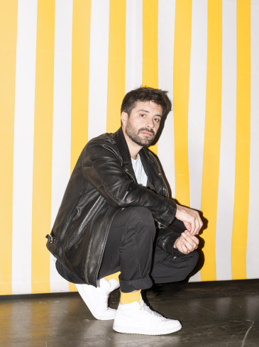 Regisseur Antú Romero Nunes trägt ein schwarzes Outfit samt Lederjacke, weiße Schuhe und hockt seitlich vor einer gelb-weiss-gestreiften Wand. 