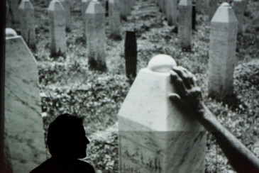 Inszenierungsfoto von Srebrenica, links im Bild die Umrisse von Jens Harzer vor einer Projektion eine Fotos im Hintergrund. Auf dem Foto sind Grabsteine in Form von Stelen zu erkennen sowie eine Hand, die einen dieser berührt. 