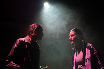 Nahaufnahme von Amal Keller und Amelie Willberg, die sich auf dunkler Bühne gegenüberstehen und anschauen. Lediglich von oben gibt es eine kleine Lichtquelle. Die Bühne ist in Nebel gehüllt.