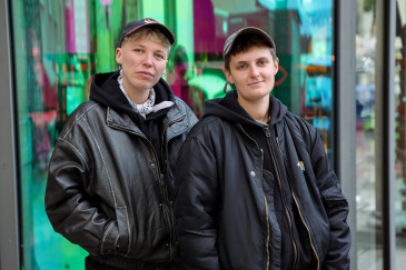 Jules Head und Leo Lorena Wyss stehen in schwarzen Bomberjacken und mit Caps vor der holografisch spiegelnden Glasfront des BE-Shops. Sie schauen ernst, stehen nah beieinander und schauen direkt in die Kamera. 