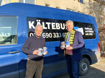 Oliver Reese übergibt dem Vorstand der Berliner Stadtmission die gesammelten Spenden. Sie stehen vor einem blauen Kältebus. Der Vorstand trägt einen Merchandiseschal des Berliner Ensembles. 